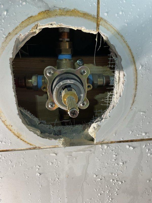 Accessed and inspected plumbing valve assembly through existing wall opening in bathroom fixture area. Valve assembly showed evidence of previous water activity with visible staining and mineral deposits on surrounding surfaces. Examined brass shut-off valve with dual blue-handled connections and verified accessibility for future maintenance requirements. External hose bib or water connection point on brick exterior was also inspected to ensure proper mounting and operational integrity of the metallic access panel and hinge assembly.
