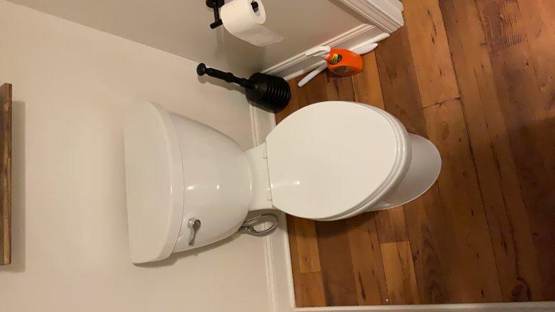Completed toilet installation in residential bathroom. Installed new white porcelain elongated bowl toilet with chrome flush handle on existing wooden plank flooring, ensuring proper positioning, secure mounting, and leak-free connections to existing plumbing.
