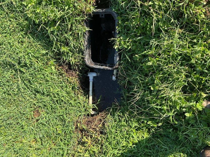 # Drainage System Access Box Inspection and Maintenance

Performed inspection and maintenance on an underground drainage system access box and associated PVC piping infrastructure. Located and accessed the ground-level junction box installed in the lawn area, which showed signs of weathering and required attention. Inspected the interior cavity of the access box for proper function and debris accumulation.

Excavated and exposed the PVC drainage piping system to assess condition and verify proper installation. Examined the T-junction configuration and elbow fittings for structural integrity, checking for cracks, leaks, or separation at connection points. The piping system showed soil accumulation and weathering consistent with ground-level installation.

Evaluated the drainage flow patterns and water accumulation in the immediate area, noting moisture levels in surrounding soil and standing water conditions. Assessed the integration point between the pipe system and landscape, ensuring