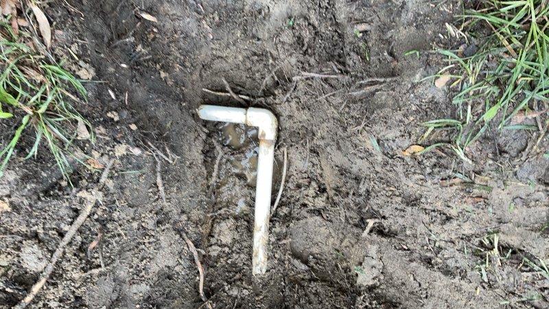 Excavated and exposed underground PVC drainage pipe for inspection and repair access. Removed soil and grass cover to locate the pipe system, revealing a right-angle pipe connection beneath the surface. Site was left prepared for subsequent plumbing repairs or modifications to the drainage infrastructure.