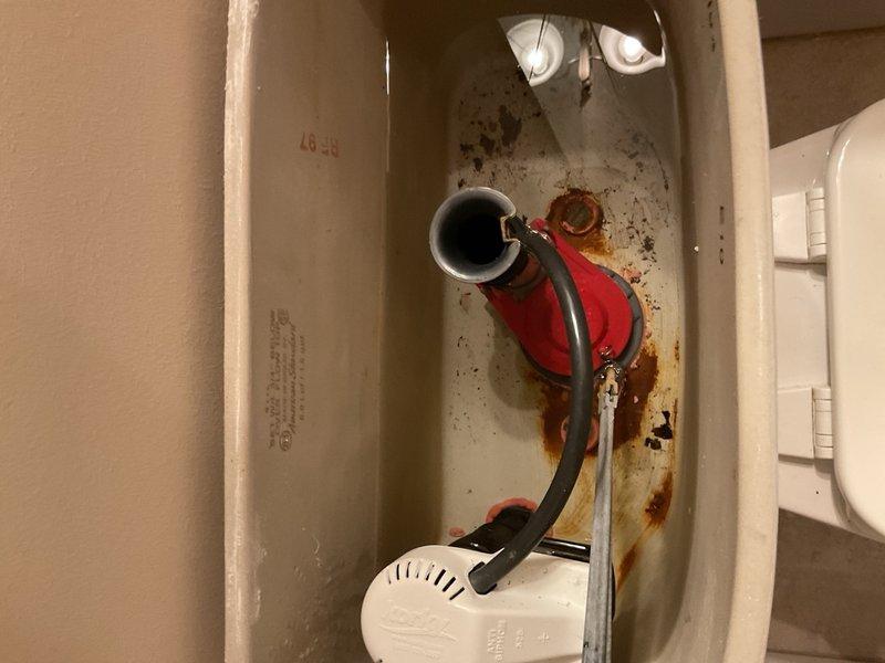 Inspected toilet tank and water supply system due to visible rust staining and corrosion buildup. Documented interior tank condition showing significant mineral deposits, rust discoloration around fill valve assembly, and deterioration of internal components. Assessed water supply line connection, fill valve mechanism with flexible hose assembly, and float mechanism for proper operation and potential replacement needs.