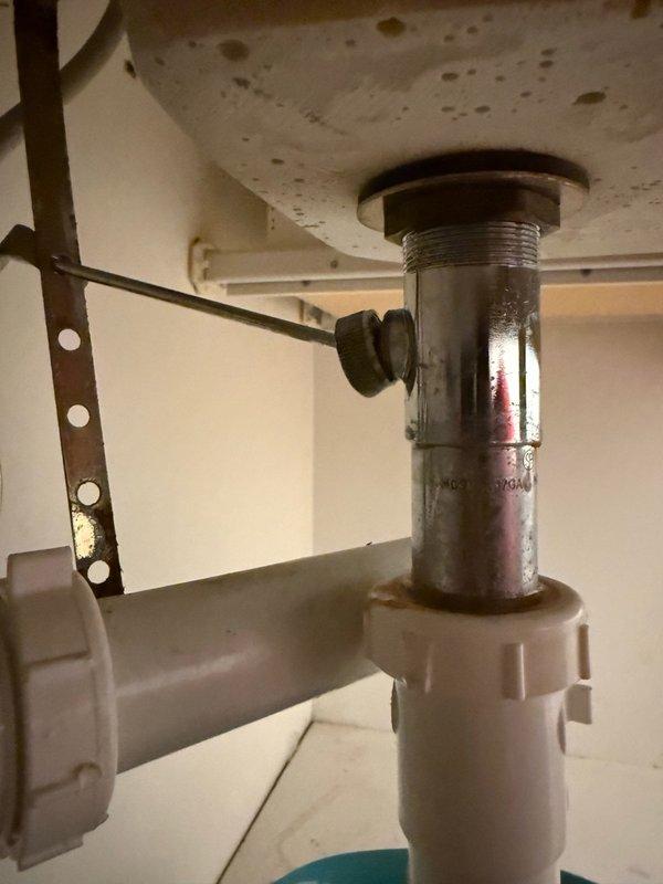 Inspected and photographed bathroom sink drain assembly and P-trap configuration. Documented existing plumbing components including chrome drain fitting, rubber gasket connections, mounting hardware, and white plastic P-trap for service records. Assessment revealed age-related discoloration and possible corrosion on metal drain surfaces requiring monitoring for future maintenance needs.
