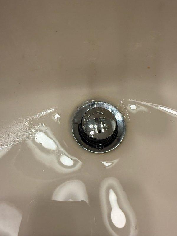 Inspected and photographed bathroom sink drain assembly and P-trap configuration. Documented existing plumbing components including chrome drain fitting, rubber gasket connections, mounting hardware, and white plastic P-trap for service records. Assessment revealed age-related discoloration and possible corrosion on metal drain surfaces requiring monitoring for future maintenance needs.
