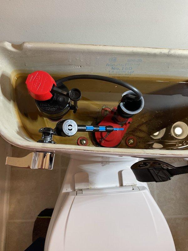 Responded to service call for toilet maintenance. Upon arrival, removed tank lid to inspect internal flush mechanism components. Found discolored water in tank indicating mineral deposits and potential supply line issues. Examined fill valve assembly with red adjustment cap, overflow tube with rubber flapper, and float mechanism. Inspected flexible water supply line connection and chrome shut-off valve at wall mounting point. Assessed Mansfield brand toilet for proper operation and identified need for water quality evaluation due to rust staining or mineral buildup in tank. Documented fixture condition and internal component status through photographs for service records.