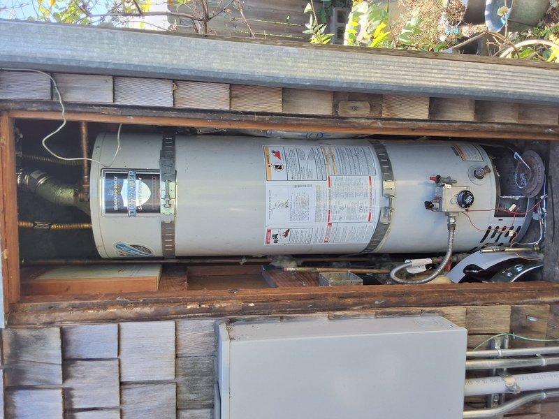 Completed residential water heater replacement with installation of a 30-gallon natural gas unit (model URG130T6N) in an exterior wooden enclosure. The installation included all necessary plumbing connections, gas line hookup with flexible yellow connector, pressure relief valve, drain pan, and proper venting. All safety labels and operational instructions are clearly visible on the unit, and the system was tested and confirmed operational upon completion.