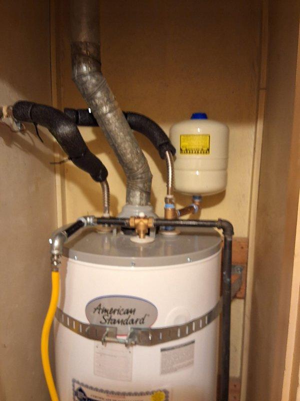 Completed warranty installation of American Standard residential water heater model GN50T1-3-6, serial number D26-017900, in utility closet enclosure. Unit was properly secured with metal safety strap, connected to copper water lines with shut-off valves, yellow gas line, expansion tank with pressure relief, and venting system with flexible ductwork. Installation included drain pan placement and verified all plumbing connections, gas fittings, and safety components met code requirements for proper operation.