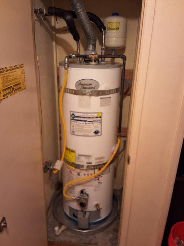 Completed warranty installation of American Standard residential water heater model GN50T1-3-6, serial number D26-017900, in utility closet enclosure. Unit was properly secured with metal safety strap, connected to copper water lines with shut-off valves, yellow gas line, expansion tank with pressure relief, and venting system with flexible ductwork. Installation included drain pan placement and verified all plumbing connections, gas fittings, and safety components met code requirements for proper operation.