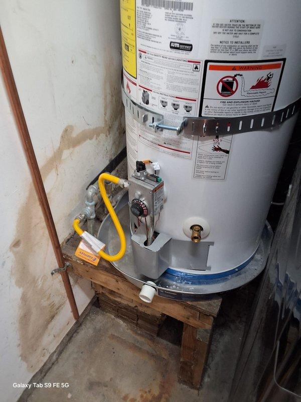 Completed no-charge warranty service on gas water heater model GN40T1-3-6, serial M25-062300, within first year of installation. Inspected unit installation, verified proper gas line connections, plumbing connections, and operational status. Documented equipment condition with photos showing water heater properly positioned on pallet base with copper piping, yellow flexible gas line, brass drain valve, and all manufacturer labels intact in utility room setting alongside stacked washer/dryer unit.