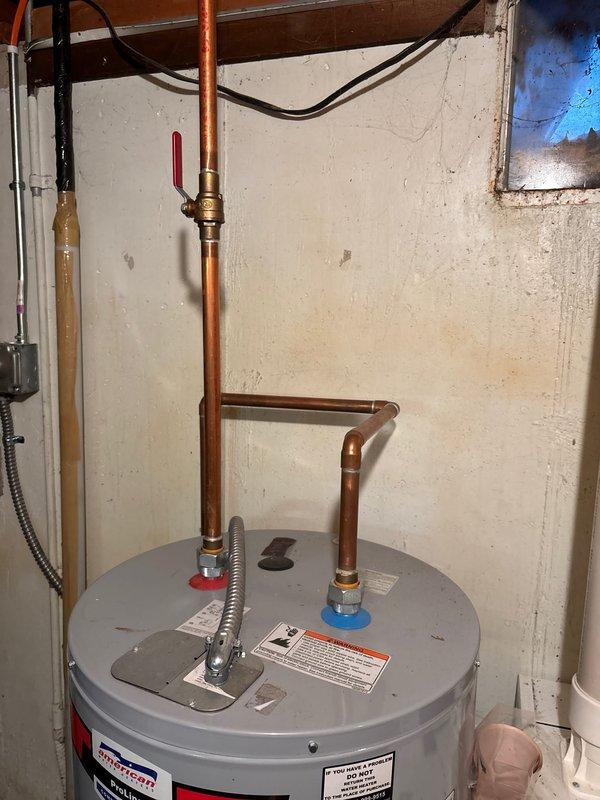 Performed comprehensive assessment of existing water heater installation in response to inadequate hot water capacity for household demand. Inspected aging water heater unit located in basement utility area, noting gray cylindrical tank with copper piping connections, brass fittings, and loop configuration at top of unit. Evaluated current system capacity relative to home age (circa 1970) and occupant usage patterns indicating hot water depletion after multiple consecutive showers. Documented equipment condition through photographic records showing water heater positioning against concrete block wall with associated plumbing infrastructure, electrical conduit connections, and visible warning labels. Observed moisture staining on concrete floor at base of unit and general signs of system age. Assessed copper supply lines, red-handled ball valve, brass fittings, and overall plumbing configuration. Reviewed manufacturer specifications on unit labeling and determined replacement recommenda