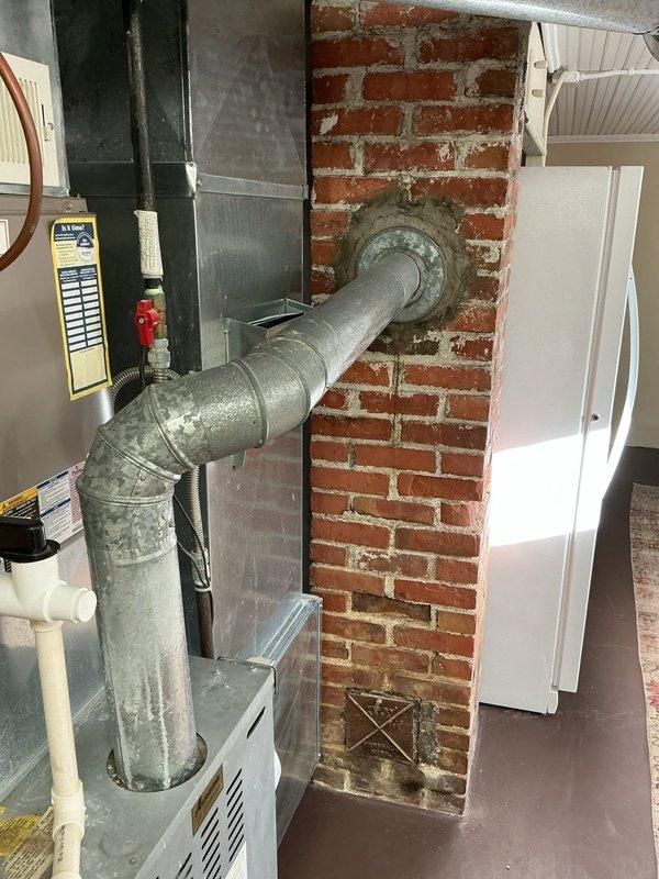 Completed estimate for full HVAC system replacement in residential basement mechanical room. Assessed existing furnace and air conditioning system located against exposed brick chimney wall with gray metal ductwork configuration. Evaluated current installation including galvanized metal supply ducts with 90-degree elbow connections, white PVC condensate drainage system, and electrical panel connections. Documented mechanical room layout featuring aged ductwork with visible surface patina and existing equipment positioning relative to brick chimney structure. Provided comprehensive quote for new furnace, air conditioning unit, and humidifier installation to replace current heating and cooling system. Photographed existing equipment setup, ductwork routing, electrical connections, and available workspace for planned installation.