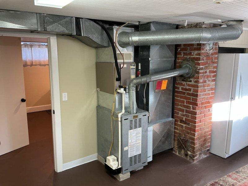 Completed estimate for full HVAC system replacement in residential basement mechanical room. Assessed existing furnace and air conditioning system located against exposed brick chimney wall with gray metal ductwork configuration. Evaluated current installation including galvanized metal supply ducts with 90-degree elbow connections, white PVC condensate drainage system, and electrical panel connections. Documented mechanical room layout featuring aged ductwork with visible surface patina and existing equipment positioning relative to brick chimney structure. Provided comprehensive quote for new furnace, air conditioning unit, and humidifier installation to replace current heating and cooling system. Photographed existing equipment setup, ductwork routing, electrical connections, and available workspace for planned installation.