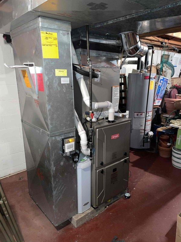 Completed full air conditioning system replacement in residential basement mechanical room. Installed new Bryant HVAC equipment including air handler unit and associated components. Ran and connected white PVC condensate drainage lines between units and routed to proper termination points. Installed refrigerant line set with copper piping, securing lines along ceiling joists to outdoor condensing unit. Mounted auxiliary equipment including humidifier or water treatment device on wall adjacent to main system. Connected all electrical wiring and control circuits to ensure proper system operation. Tested system startup, verified airflow through ductwork, and confirmed all safety controls functioning correctly. Left equipment operational and mechanical room secure.