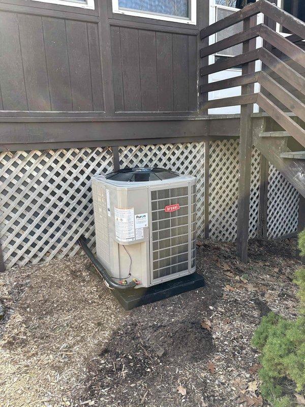 Completed full air conditioning system installation including Bryant 16.5 SEER2 condenser unit positioned on new protective pad in ground-level area beneath elevated deck. Installed new line set with proper refrigerant charge of 5.5 lbs initial plus 11 oz added charge per manufacturer specifications. Unit mounted on black equipment pad over wood chip base with lattice skirting, all connections secured and system tested for proper operation.