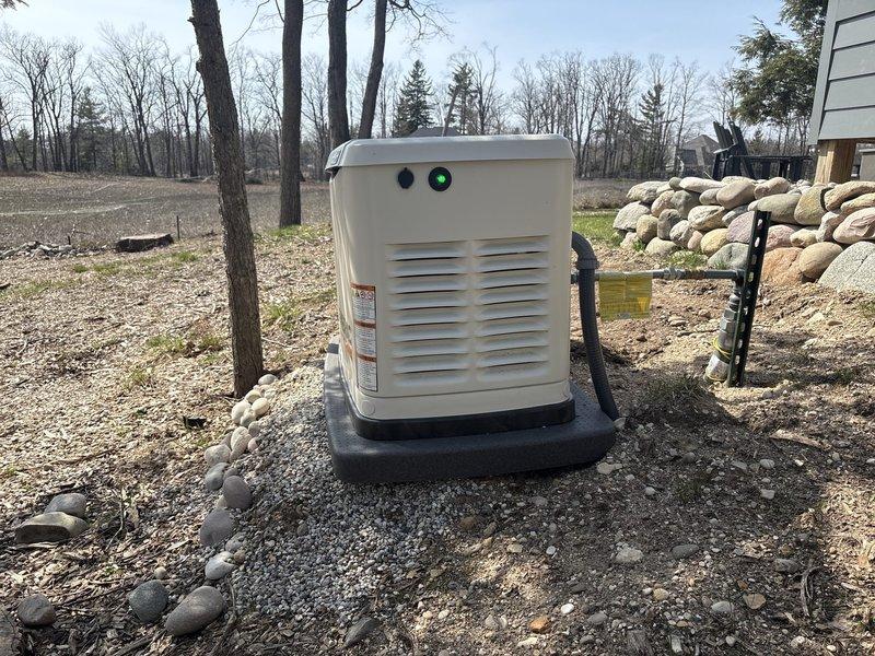 Returned to residential generator installation site for spring follow-up inspection. Verified trenching work from initial installation was properly backfilled and graded. Inspected decorative stone retaining wall adjacent to generator pad to confirm structural integrity and proper retention. Documented generator unit condition and surrounding landscape with photos showing concrete pad, gravel base, and rock wall all in satisfactory condition.