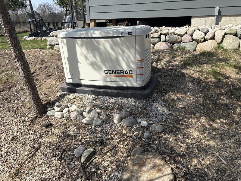 Returned to residential generator installation site for spring follow-up inspection. Verified trenching work from initial installation was properly backfilled and graded. Inspected decorative stone retaining wall adjacent to generator pad to confirm structural integrity and proper retention. Documented generator unit condition and surrounding landscape with photos showing concrete pad, gravel base, and rock wall all in satisfactory condition.