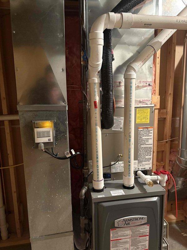 Completed full HVAC system replacement including outdoor condensing unit and indoor air handler components. Installed new Armstrong Air furnace system in basement mechanical room with properly configured PVC exhaust and intake venting. Positioned gray cylindrical outdoor AC condenser unit on concrete pad adjacent to residential structure with adequate clearance from building siding and nearby stairway. Connected all refrigerant lines, electrical components, and drainage systems per manufacturer specifications. Verified proper operation of both indoor and outdoor equipment, confirmed airflow through ductwork, and ensured all safety labels and warning placards remain visible and intact.