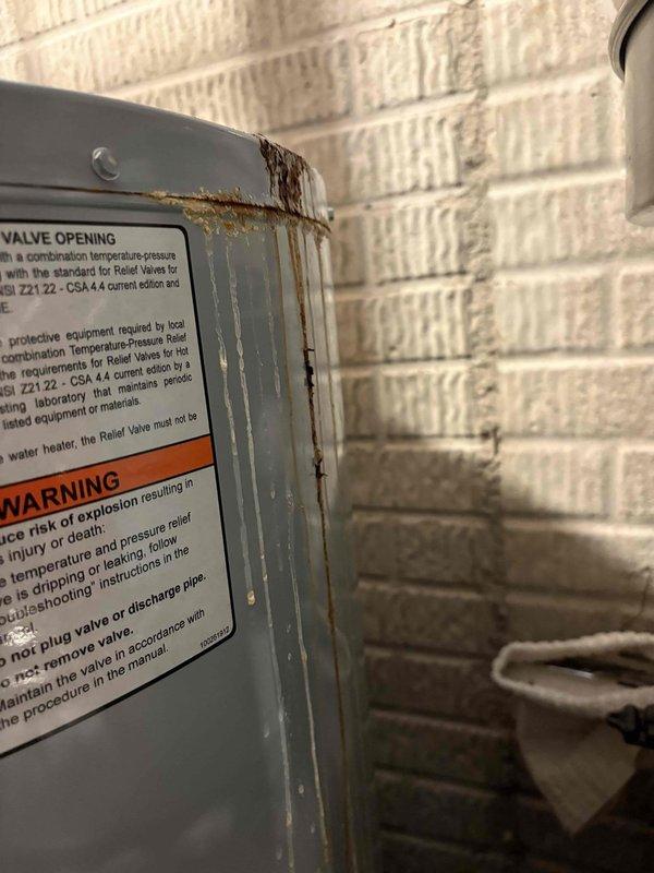 Responded to water heater leak in drain pan. Upon inspection, found significant corrosion and rust accumulation at copper pipe union connection above unit, with evidence of active water seepage causing rust staining on surrounding surfaces. Verified unit is a 4-year-old American-brand water heater showing advanced corrosion on exterior panels and pipe connections. Documented extensive rust streaking on unit casing and deteriorated pipe fittings. Verified warranty coverage and photographed conditions for warranty claim processing.