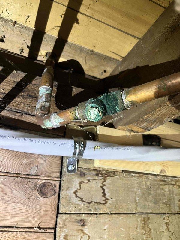 # Plumbing Leak Repair - Main Level Tub and Toilet Supply Valve

Responded to report of main level bathtub leaking into basement and toilet supply valve leak in blue bathroom. Upon arrival, accessed property through unlocked front door as instructed.

Inspected basement area beneath main level tub and identified active leak dripping through floor joists. Found makeshift towel placed under leak point to catch water. Traced leak source to deteriorated drain assembly connections above. Examined exposed copper supply lines and drain piping in basement ceiling, noting significant corrosion and verdigris buildup on copper pipes at joint connections. Documented extensive oxidation on aged copper piping and fittings, indicating long-term moisture exposure and material degradation.

Inspected blue bathroom toilet supply valve and confirmed active leak at valve connection point. Evaluated condition of existing shut-off valve and supply line. Assessed surrounding plumbing infrastructure including