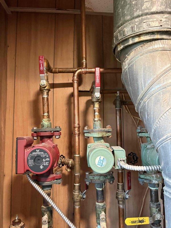 Assessed existing hydronic heating system for potential removal of baseboard heating in one room. Inspected dual circulation pump configuration with red and green circulators connected via copper piping with isolation valves, documenting current system layout and zone control setup. Evaluated floor joist access points and examined structural considerations for baseboard removal and potential pipe modifications in the affected area.
