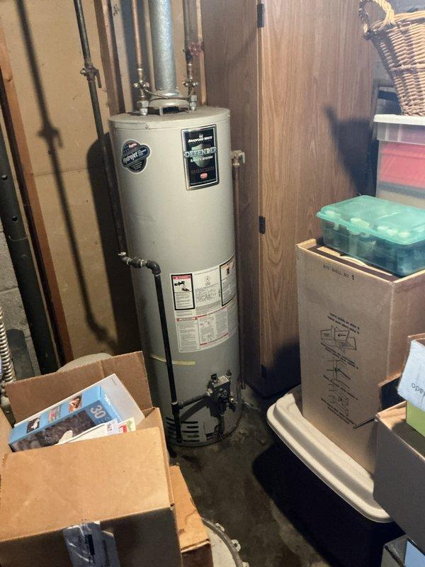Responded to customer report of leaking water heater in basement utility area. Upon inspection, observed water pooling at base of white cylindrical water heater unit with visible moisture accumulation on floor. Documented existing equipment configuration including galvanized vent piping, multiple connection points at top of unit, and current installation against wood-paneled walls in cluttered storage space. Photographed water heater specifications, surrounding plumbing infrastructure including insulated pipes, and basement mechanical room conditions to provide comprehensive estimate for water heater replacement.