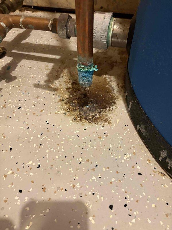 Responded to water leak call and conducted diagnostic inspection. Upon arrival, identified source of leak as corroded temperature and pressure relief valve discharge pipe on water heater. Observed significant turquoise-green copper corrosion at pipe terminus with evidence of water damage on floor surface beneath. Documented conditions with photos showing advanced oxidation of copper piping and brownish residue staining on utility room floor. Advised homeowner of findings and recommended replacement of corroded discharge piping to prevent continued leakage.