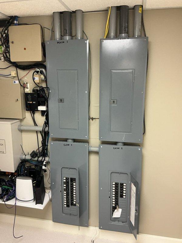 Completed comprehensive site evaluation for standby generator installation. Assessed existing electrical infrastructure including dual main service panels (MAIN 1, GEN 1, and GEN 2 sections) with exposed circuit breaker configurations. Documented current automatic transfer switch installation rated for NEMA 3R indoor/outdoor use, verifying compatibility with proposed generator system. Evaluated existing conduit pathways including gray PVC and yellow conduit runs from panel tops, noting horizontal white PVC interconnections between panel systems. Photographed auxiliary electrical components including wall-mounted controller with indicator lights, surge protection equipment, and associated black cable routing throughout the installation area. Reviewed networking and communications equipment positioning near transfer switch to ensure clearance requirements for generator integration. Assessed overall electrical distribution system capacity and documented all relevant specifications for sta