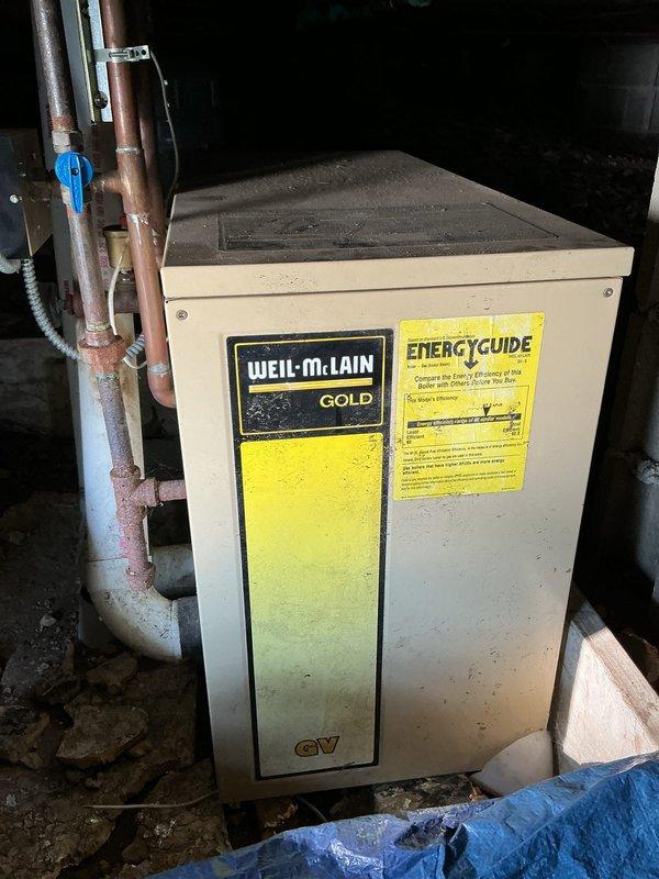 Conducted on-site assessment for replacement of 30-year-old heating system. Upon inspection, identified existing equipment as a Weil-McLain Gold boiler unit showing significant age-related wear, dust accumulation, and surface discoloration. Evaluated copper supply and return piping displaying oxidation and patina consistent with system age. Documented current installation configuration including piping connections, electrical routing, and equipment placement in basement utility area. Assessed crawl space conditions revealing aged copper distribution lines with corrosion, electrical wiring runs along floor joists, and concrete block foundation supports. Captured photographic documentation of existing boiler unit, associated plumbing connections, and underfloor distribution system for replacement planning purposes. Provided comprehensive evaluation for complete heating system replacement estimate.