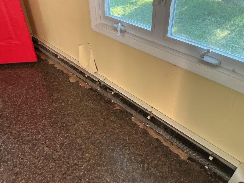 Consulted with contractor regarding baseboard kick plate installation in multi-room residential space. Documented existing baseboard heating units along yellow and mint green painted walls, noting heater cover requiring proper securing and assessing transition areas between carpeted and hardwood flooring for kick plate installation specifications.