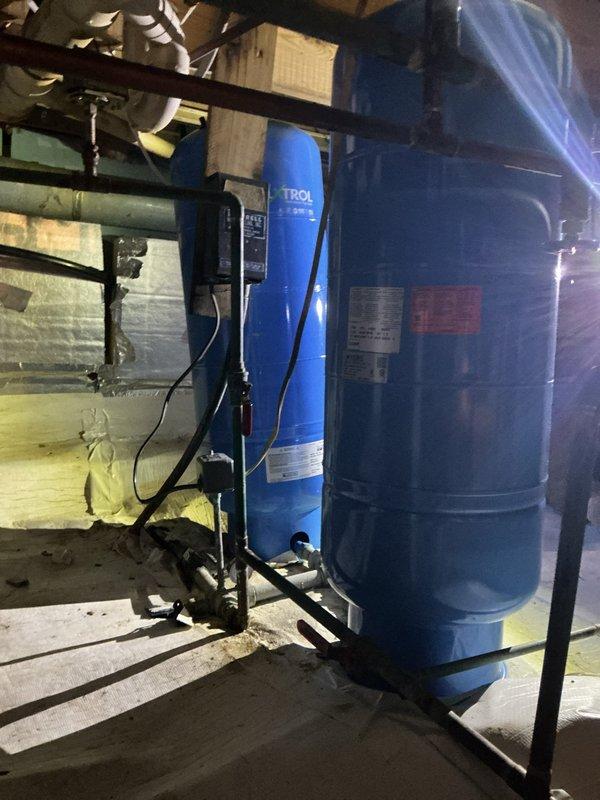 Responded to low water pressure and insufficient hot water throughout 40-year-old residence. Upon inspection, evaluated existing water pressure tank system and water treatment equipment in basement mechanical room. Documented current configuration of blue TROL-branded pressure tank and accompanying dark storage tank, both professionally mounted on black metal frame with proper electrical controls and piping connections. System components showed age-appropriate wear consistent with original home construction. Photographed installation for service records, including all visible piping connections, electrical conduit, control box, and mounting hardware to establish baseline for future maintenance recommendations.