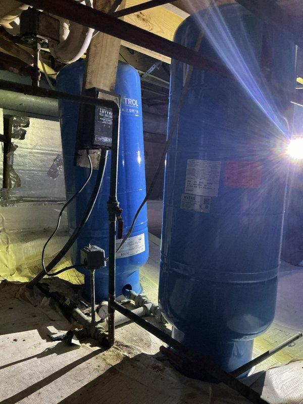 Responded to low water pressure and insufficient hot water throughout 40-year-old residence. Upon inspection, evaluated existing water pressure tank system and water treatment equipment in basement mechanical room. Documented current configuration of blue TROL-branded pressure tank and accompanying dark storage tank, both professionally mounted on black metal frame with proper electrical controls and piping connections. System components showed age-appropriate wear consistent with original home construction. Photographed installation for service records, including all visible piping connections, electrical conduit, control box, and mounting hardware to establish baseline for future maintenance recommendations.