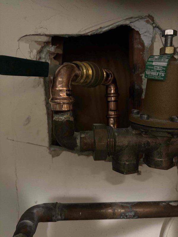 Inspected leaking water main shut off valve. Accessed plumbing system through wall opening, revealing copper pipes with visible corrosion and a brass pressure regulator valve with green tag. Identified shut off valve with red handle as source of leak when operated. Inspected leaking water main shut off valve. Accessed plumbing system through wall opening, revealing copper pipes with visible corrosion and a brass pressure regulator valve with green tag. Identified shut off valve with red handle as source of leak when operated.