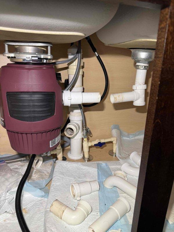 Installed customer-supplied garbage disposal (burgundy/maroon model) under sink and corrected existing plumbing configuration. Completed proper connections to drain system using white PVC piping with appropriate joints and elbows, ensuring secure attachment to sink baskets above. Verified electrical connections and performed operational test confirming proper drainage and disposal function. Installed customer-supplied garbage disposal (burgundy/maroon model) under sink and corrected existing plumbing configuration. Completed proper connections to drain system using white PVC piping with appropriate joints and elbows, ensuring secure attachment to sink baskets above. Verified electrical connections and performed operational test confirming proper drainage and disposal function.