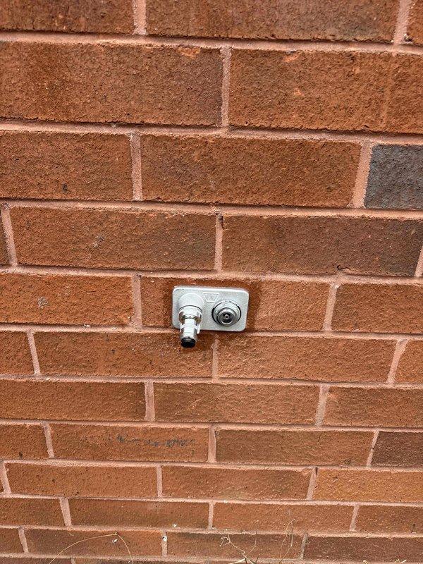 Inspected exterior brick wall water spigot fixture and interior commercial utility room. Documented condition of wall-mounted water connection point with metallic base plate and examined interior mechanical space containing exposed ductwork, electrical conduits, junction boxes, and service panel for approved quote specifications. Inspected exterior brick wall water spigot fixture and interior commercial utility room. Documented condition of wall-mounted water connection point with metallic base plate and examined interior mechanical space containing exposed ductwork, electrical conduits, junction boxes, and service panel for approved quote specifications.