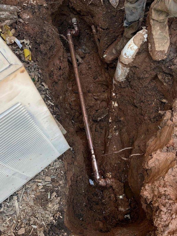Excavated and exposed underground copper piping system for inspection and repair access. Documented existing plumbing infrastructure including oxidized copper pipe with branch connections and adjacent concrete utility components positioned near HVAC equipment foundation. Completed trenching work and site photography for service records. Excavated and exposed underground copper piping system for inspection and repair access. Documented existing plumbing infrastructure including oxidized copper pipe with branch connections and adjacent concrete utility components positioned near HVAC equipment foundation. Completed trenching work and site photography for service records.