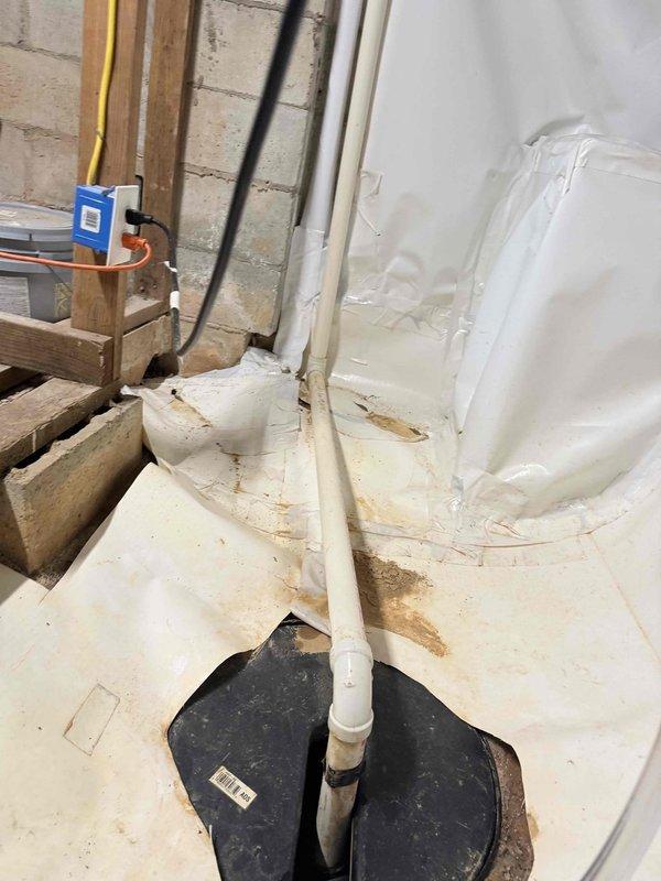 Responded to service call for sump pump inspection with concerns regarding discharge piping functionality. Upon arrival, evaluated exterior discharge line termination point at foundation wall and confirmed proper outlet configuration through foundation vent area. Inspected interior sump basin installation, including vertical discharge pipe with floor penetration boot assembly. Observed evidence of previous moisture infiltration around pipe penetration point based on staining patterns on protective floor sheeting. Verified discharge pipe routing, connections, and mechanical systems integration. Documented current system condition with photographic evidence for service records. Responded to service call for sump pump inspection with concerns regarding discharge piping functionality. Upon arrival, evaluated exterior discharge line termination point at foundation wall and confirmed proper outlet configuration through foundation vent area. Inspected interior sump basin installation, including vertical discharge pipe with floor penetration boot assembly. Observed evidence of previous moisture infiltration around pipe penetration point based on staining patterns on protective floor sheeting. Verified discharge pipe routing, connections, and mechanical systems integration. Documented current system condition with photographic evidence for service records.