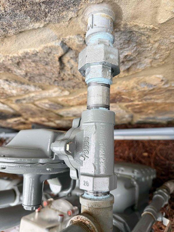 Completed installation and inspection of gas line infrastructure including vertical pipe assembly with T-junction valve and horizontal distribution lines. Installed Honeywell gas pressure regulator on exterior stone wall with proper mounting and pipe connections. All threaded fittings, couplings, and connections were secured and tested for proper operation and safety compliance.