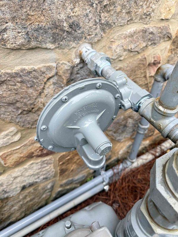 Completed installation and inspection of gas line infrastructure including vertical pipe assembly with T-junction valve and horizontal distribution lines. Installed Honeywell gas pressure regulator on exterior stone wall with proper mounting and pipe connections. All threaded fittings, couplings, and connections were secured and tested for proper operation and safety compliance.