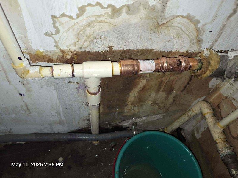 Responded to service call for leaking water heater in basement utility area. Upon inspection, identified active water leak originating from corroded connection points on aging copper supply lines. The existing plumbing infrastructure showed significant deterioration with heavy oxidation and greenish patina on copper piping, along with evidence of previous moisture intrusion on surrounding walls. Assessed transition points between newer yellow plastic piping and older metal sections, which exhibited compromised integrity. Isolated water supply to prevent further leakage and water damage. Documented extensive corrosion on horizontal pipe assembly and deteriorated connections requiring replacement. Noted compromised plumbing components with visible corrosion, tape repairs, and oxidized surfaces indicating end of service life. Recommended complete replacement of corroded copper sections and upgrading connection points to prevent future failures. Responded to service call for leaking water heater in basement utility area. Upon inspection, identified active water leak originating from corroded connection points on aging copper supply lines. The existing plumbing infrastructure showed significant deterioration with heavy oxidation and greenish patina on copper piping, along with evidence of previous moisture intrusion on surrounding walls. Assessed transition points between newer yellow plastic piping and older metal sections, which exhibited compromised integrity. Isolated water supply to prevent further leakage and water damage. Documented extensive corrosion on horizontal pipe assembly and deteriorated connections requiring replacement. Noted compromised plumbing components with visible corrosion, tape repairs, and oxidized surfaces indicating end of service life. Recommended complete replacement of corroded copper sections and upgrading connection points to prevent future failures.