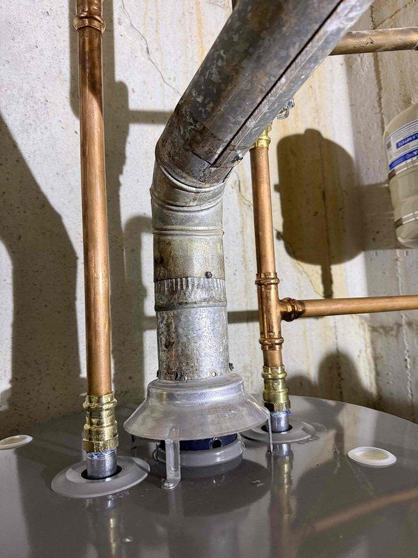 Responded to customer complaint of gas or burning odor near water heater. Performed thorough inspection of water heater and surrounding plumbing components. Examined copper supply lines, brass fittings, and galvanized flue vent pipe for signs of gas leaks or combustion issues. Documented heavily corroded and deteriorated flue vent system with significant mineral buildup and corrosion on metal surfaces. Identified crusty white and yellowish deposits indicating potential exhaust condensation issues and compromised venting. Photographed installation showing aged galvanized flue pipe with visible weathering and HVAC components with substantial mineral accumulation. Recommended immediate evaluation of venting system due to corrosion severity and potential safety concerns related to improper exhaust ventilation.