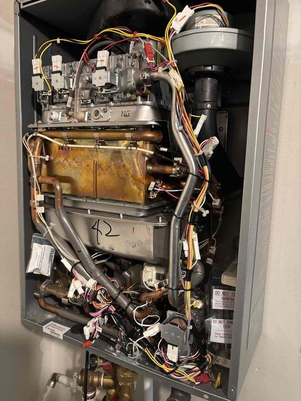 Responded to no hot water complaint on Rinnai tankless water heater. Upon inspection, found unit powered on with controller displaying 130°F setpoint but not producing hot water at fixtures. Removed front panel and performed diagnostic inspection of internal components. Identified significant corrosion and oxidation on heat exchanger assembly, indicating potential scale buildup or component failure affecting heat transfer capability. Checked gas valve assemblies, blower motor operation, and electrical connections throughout the unit. Unit was not displaying error codes despite operational failure, suggesting possible sensor malfunction or communication issue between components and controller. Documented current conditions with photos showing corroded heat exchanger and control panel status for further evaluation and repair recommendations.