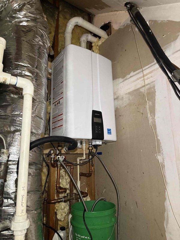 Responded to service call for tankless water heater making loud noise and toilet flush issue. Inspected recently installed tankless unit with complete plumbing assembly including pressure relief system, isolation valves, and pressure gauge. Performed system flush on tankless water heater to address noise concerns and verified proper operation of all connected components including blue-handled isolation valves and pressure regulation equipment. Documented installation configuration showing proper mounting, venting through metallic flex ductwork, and appropriate safety equipment including collection bucket beneath unit.