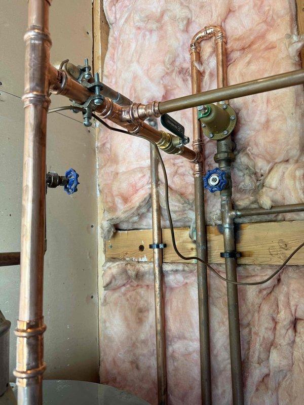 Completed thermal expansion tank rework and main shut-off valve installation in mechanical room. Reconfigured copper piping system with new ball valves for improved isolation and system control. Installed white expansion tank with secure mounting brackets to accommodate thermal expansion in the water heating system. All connections properly soldered and tested for leaks, with multiple shut-off points strategically placed throughout the piping network for future maintenance accessibility.