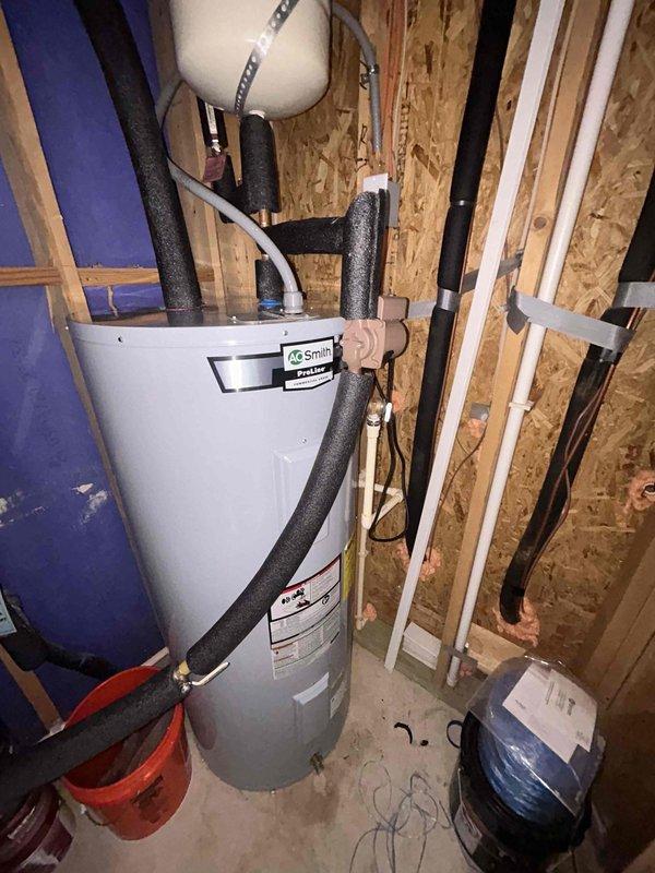 Replaced water heater and associated plumbing connections in mechanical room. Installed new white cylindrical water heater with proper pipe insulation on copper supply lines. Connected flexible metallic tubing and integrated unit with existing blue pressure tank system. Cleaned and serviced brass valve component that showed significant internal sediment buildup and deposits. Verified all connections for proper operation and system integrity.
