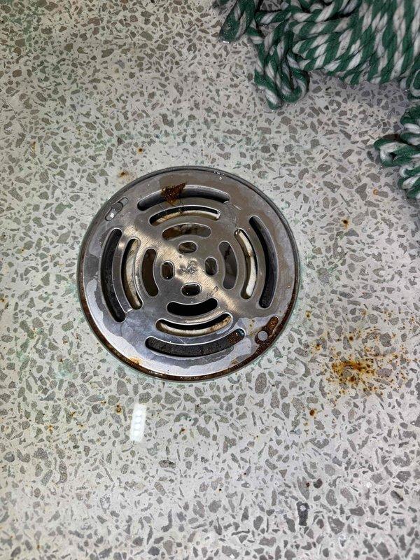 Responded to service call for drainage issue with uncertain origin between interior drain line and exterior gutter system. Upon inspection, identified a floor drain with circular metal grate showing signs of age and potential flow restriction in what appears to be a bathroom or wet room area. Traced drainage path to exterior where downspout terminates at building foundation, discovering significant water pooling and moisture accumulation at the base indicating improper drainage or blockage in the exterior discharge system. Documented conditions of both interior drain fixture and exterior downspout outlet to determine source of clogging issue.
