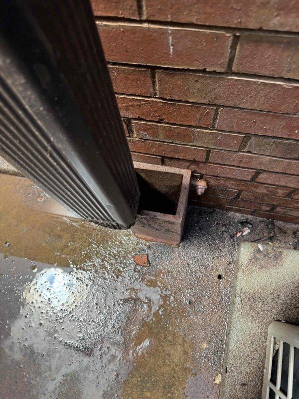 Responded to service call for drainage issue with uncertain origin between interior drain line and exterior gutter system. Upon inspection, identified a floor drain with circular metal grate showing signs of age and potential flow restriction in what appears to be a bathroom or wet room area. Traced drainage path to exterior where downspout terminates at building foundation, discovering significant water pooling and moisture accumulation at the base indicating improper drainage or blockage in the exterior discharge system. Documented conditions of both interior drain fixture and exterior downspout outlet to determine source of clogging issue.