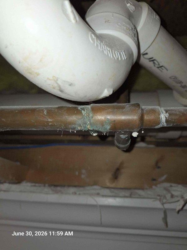 Responded to leak complaint with water penetrating ceiling from bathroom above. Upon inspection, identified active leak on corroded copper supply line beneath bathroom fixture. Copper piping exhibited significant verdigris corrosion at joint connection with visible water droplets present. Documented existing conditions including compromised pipe integrity and moisture damage. Verified system pressure at 0 PSI on gauge, indicating supply isolation. Assessed drainage assembly including P-trap and PVC connections for additional concerns. Photographed corroded copper line, pressure gauge readings, and affected plumbing components for documentation purposes.