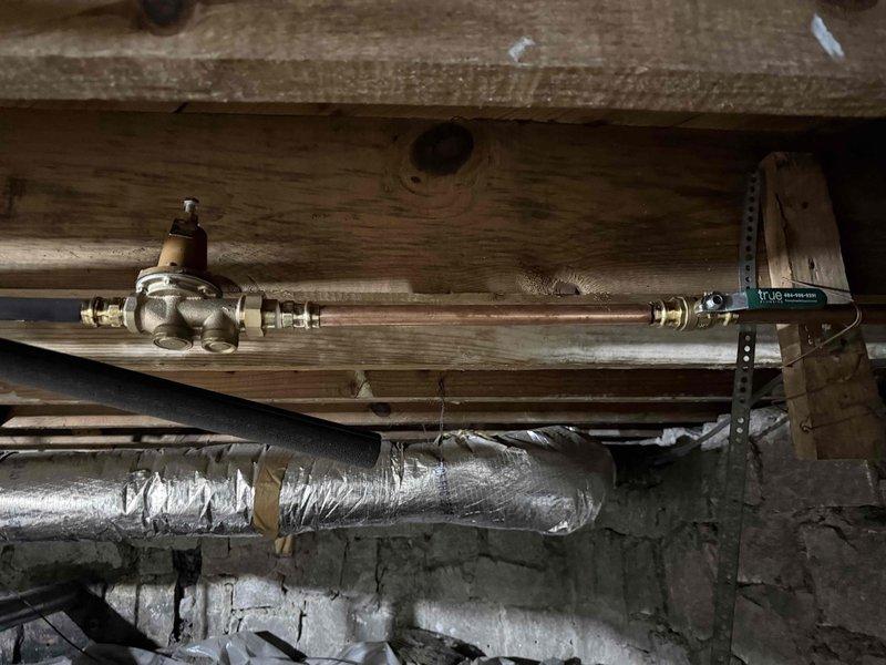 Completed follow-up service call to address high water pressure concerns following recent water service repair. Inspected and photographed the existing pressure reducing valve installation located in the crawlspace beneath the structure. The brass pressure reducing valve with adjustment mechanism was found properly installed on the copper water line with secure compression fittings on both inlet and outlet connections. Valve was mounted to floor joists using appropriate support strapping to prevent stress on pipe connections. All components appeared to be functioning as designed with no visible leaks or defects noted at the time of inspection.