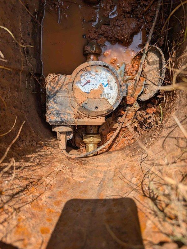 Completed site assessment and documentation of water meter assembly requiring replacement. Photographed heavily corroded meter installation showing significant rust deterioration, root intrusion, and accumulated debris in underground utility vault. Documented roadside drainage area with municipal grate system adjacent to paved surface. Utility locate request submitted per 811 requirements prior to excavation work.