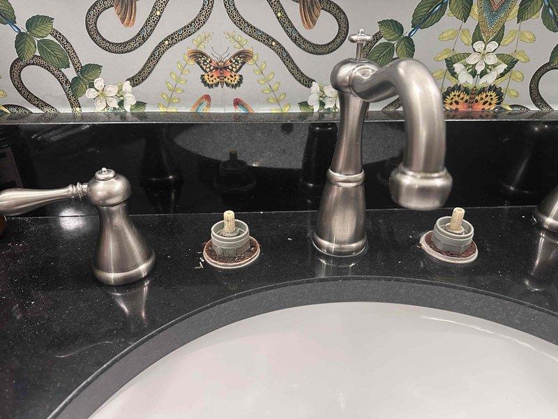Responded to service call for leaking bathroom faucet with three-handle configuration (hot/cold handles and center spout) in brushed nickel finish mounted on black granite countertop. Upon inspection, identified deteriorated seals and gaskets at base of fixture causing water seepage, evidenced by mineral deposits and buildup around mounting plates. Provided comprehensive assessment and repair estimate for seal replacement and proper reseating of faucet components to eliminate leak. Responded to service call for leaking bathroom faucet with three-handle configuration (hot/cold handles and center spout) in brushed nickel finish mounted on black granite countertop. Upon inspection, identified deteriorated seals and gaskets at base of fixture causing water seepage, evidenced by mineral deposits and buildup around mounting plates. Provided comprehensive assessment and repair estimate for seal replacement and proper reseating of faucet components to eliminate leak.