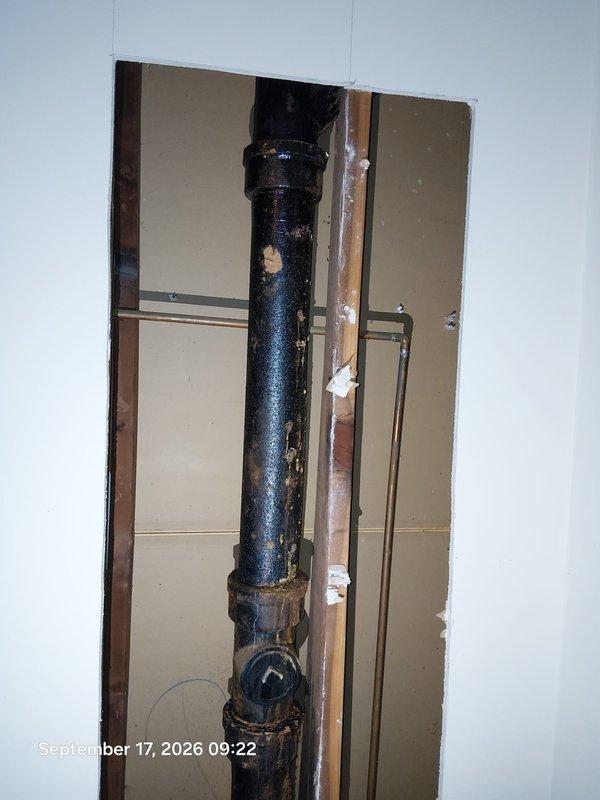 Responded to bathroom leak complaint and performed diagnostic inspection. Opened interior wall to expose plumbing infrastructure and identified deteriorated vertical waste pipe with significant corrosion and surface degradation at multiple coupling joints. Black drain pipe exhibited rust, discoloration, and aging throughout its vertical run, with visible moisture staining on surrounding floor surfaces indicating active or recent water infiltration. Documented condition of compromised pipe system, exposed wall cavity revealing wooden framing and drywall backing, and adjacent copper supply line. Inspection confirmed structural pipe failure requiring replacement to prevent continued water damage. Responded to bathroom leak complaint and performed diagnostic inspection. Opened interior wall to expose plumbing infrastructure and identified deteriorated vertical waste pipe with significant corrosion and surface degradation at multiple coupling joints. Black drain pipe exhibited rust, discoloration, and aging throughout its vertical run, with visible moisture staining on surrounding floor surfaces indicating active or recent water infiltration. Documented condition of compromised pipe system, exposed wall cavity revealing wooden framing and drywall backing, and adjacent copper supply line. Inspection confirmed structural pipe failure requiring replacement to prevent continued water damage.